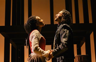 With the shadowed black outline of the wood superstructure behind them, Agnes in Simple Elizabethan dress and William in Elizabehtn leather jacket hold hands. He's looking up to the heavens, she's looking up toward his eyes.