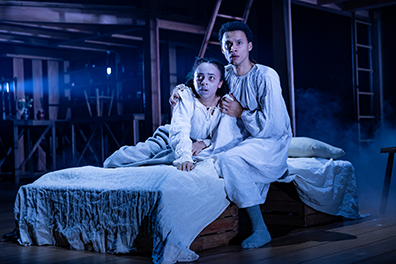 Hamnet in a white nightgown sits on the bed with one stocking foot on the floor. His hands are gripping Judith's shoulders. She's sitting up, also in white nightgown, with the bed covers over her legs. They are both looking with alarm at something past the camera's view. In the background is the wood superstructure with housewares on shelves