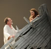 Miranda in the bow of a wrecked boat looks back, smiling, at Prospero behind her in the bow and wearing a ragged white shirt.