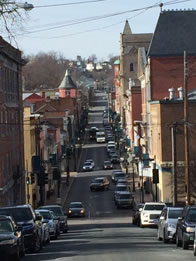 A busy two lane street down a hill and up another with a variety of old buildings on either side